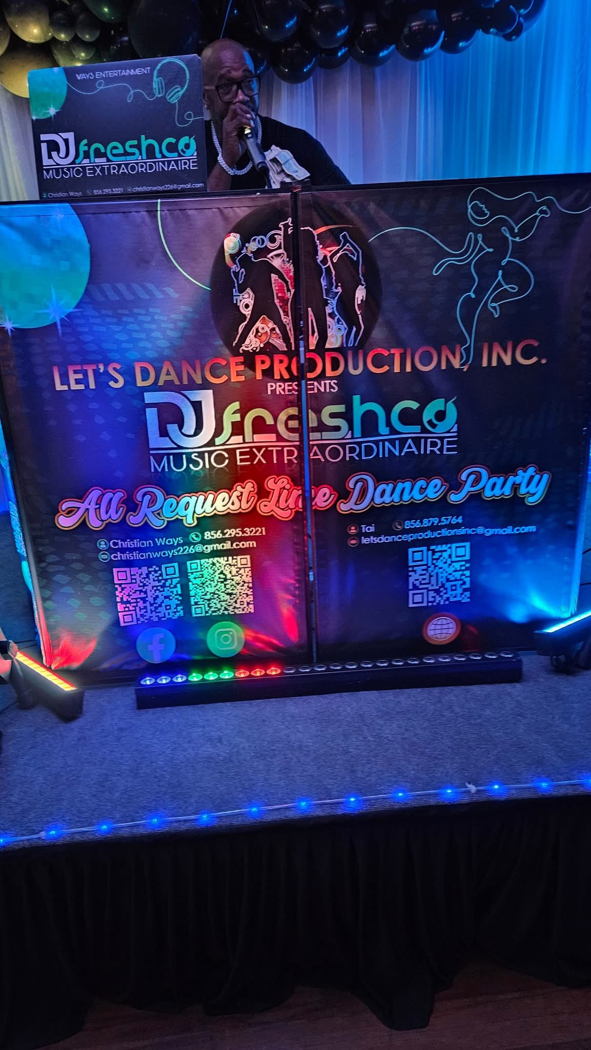 DJ Freshco holding a microphone behind his illuminated DJ booth